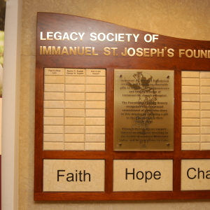 Donor recognition display for the donors of Legacy Society of Immanuel St. Joseph's Foundation. Engraved Corian tiles are surrounded by cherry stained wood with the statement "Faith, Hope, Charity"